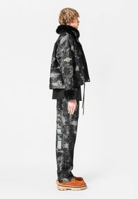 Chaqueta de denim negro con cuello de piel, textura con patrón. Pantalones negros con acentos estampados. Zapatos marrones con suela gruesa. Vista lateral.