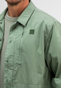 Hellgrüne Jacke aus glattem Stoff mit Kragen, Druckknöpfen und einer mit Reißverschluss versehenen Tasche. Verfügt über ein grünes, gesticktes Logo auf der Brust.