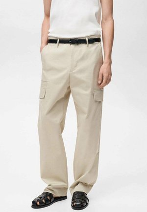 Person trägt beige Cargohose mit Seitentaschen, schwarzen Gürtel, weißes ärmelloses Oberteil und schwarze geflochtene Ledersandalen.
