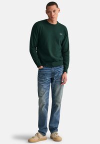Maglione verde a girocollo fatto di un tessuto morbido, con un piccolo logo. Abbinato a jeans azzurri chiari e sneaker beige.