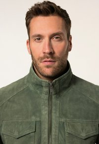 Grüne Wildlederjacke mit hohem Kragen, frontalem Reißverschluss und zwei Brusttaschen. Verfügt über eine glatte Textur und detaillierte Nähte.