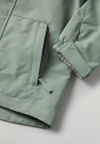 Vihreä, vettä hylkivä takki, jossa on etutasku ja joustavat hihansuut. Kankaan pinta on sileä ja siinä on hienovaraista kiiltoa.