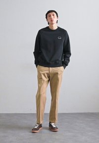 Svart hettegenser med smilende detalj, beige bukser og brune sneakers med hvite snører. Uformell antrekk på en ensfarget bakgrunn.