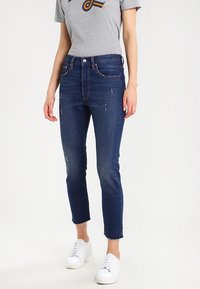 Strakzittende donkerblauwe jeans met verweerde details, vijf zakken, een knoopsluiting en rafelige zomen, gecombineerd met een grijs T-shirt en witte sneakers.