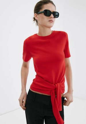 Donna con occhiali da sole neri e top rosso aderente a maniche corte con nodo annodato in vita, abbinato a pantaloni neri.