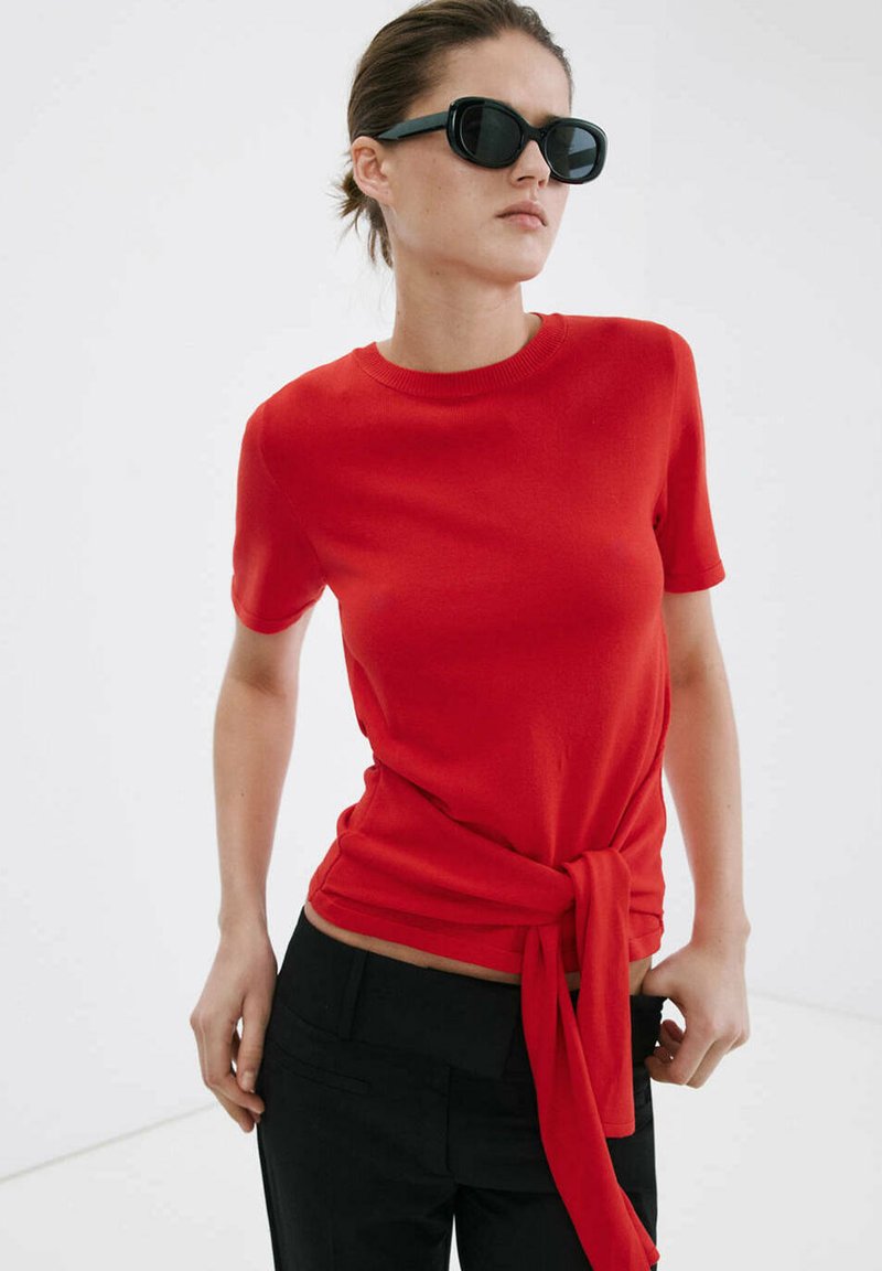 Femme portant des lunettes de soleil noires et un haut rouge ajusté à manches courtes avec un nœud noué à la taille, associé à un pantalon noir.