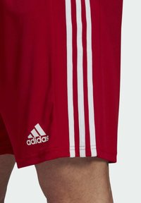 Red athletic shorts made of a smooth fabric, featuring three white stripes running down the sides and a small white logo on the hem.