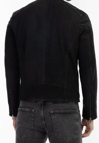 Person, der eine schwarze Lederjacke mit Reißverschlussdetails an den Ärmeln und aufgehellten schwarzen Jeans mit sichtbaren Gesäßtaschen trägt, von hinten gezeigt.