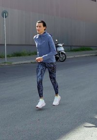 Urheiluasu, jossa on vaaleansininen vetoketjullinen takki ja kuvioidut mustat legginsit. Valkoiset sneakersit, joissa on punaisia yksityiskohtia. Kaupunkimainen katutausta.