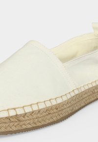 Chaussure espadrille en toile blanche avec semelle en jute tressée et design sans lacets sur un fond clair uni.