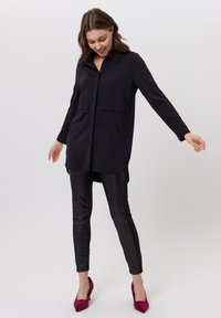 Schwarzes Langarmhemd mit durchgehender Knopfleiste und Kragen, kombiniert mit glänzenden schwarzen Leggings und fuchsiafarbenen High Heels.