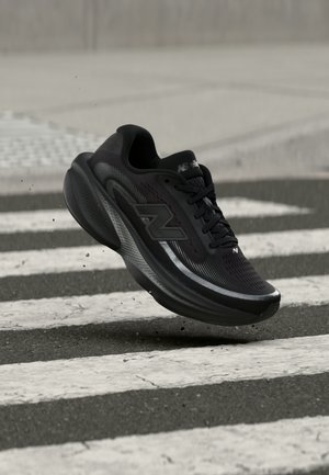 Μαύρο παπούτσι τρεξίματος New Balance με παχιά σόλα, εμφανιζόμενο στον αέρα πάνω από μια διάβαση πεζών σε έναν ασφαλτοστρωμένο δρόμο.