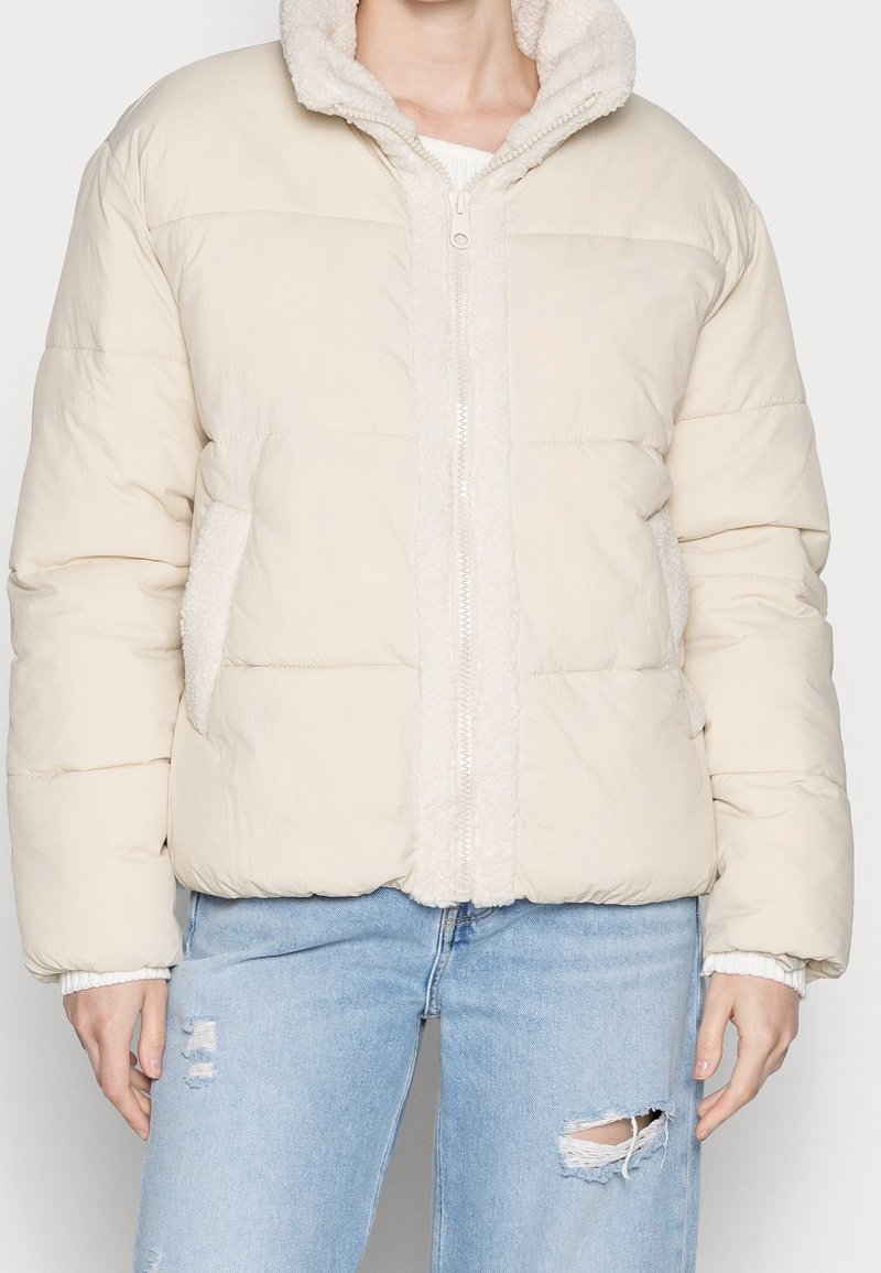 Beige pufferjakke med en tekstureret, fuzzy fór, lynlåsåbning og sidelommer. Har en let oversized pasform og pufærmer.