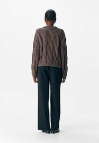 Donna in piedi con le spalle alla fotocamera, che indossa un maglione spesso a trecce marrone, pantaloni neri a gamba larga e scarpe nere.
