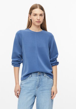 Junge Frau mit glatten Haaren, die ein blaues Langarmshirt und hellblaue Jeans trägt, steht vor einem schlichten weißen Hintergrund.