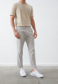 Beige crew neck t-shirt with small black logo, paired with light gray tapered pants and white sneakers with navy accent. Smooth fabric texture.