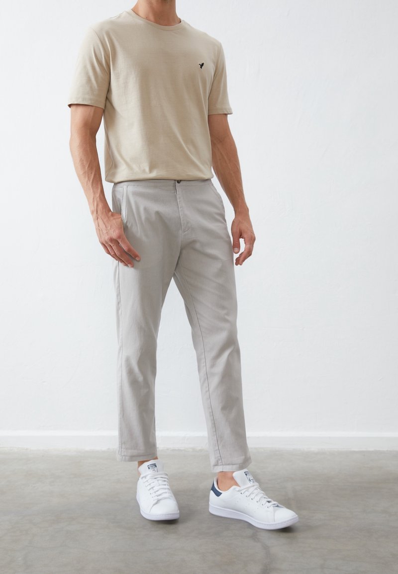 Beige crew neck t-shirt with small black logo, paired with light gray tapered pants and white sneakers with navy accent. Smooth fabric texture.