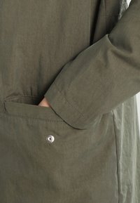 Detail lehkého olivově zeleného bundy, která ukazuje kapsu se stříbrným cvokem a hladkou, matnou texturou látky.