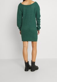 Robe pull en laine verte avec un design épaule dénudée, texture côtelée et manches longues, arrivant à mi-cuisse, associée à des bottes de combat noires.