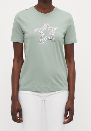 Osoba ubrana w miętowo-zieloną koszulkę z krótkim rękawem z wzorem srebrnej gwiazdy wykonanej z cekinów na przodzie, w połączeniu z białymi spodniami, stojąca na jednolitym tle.