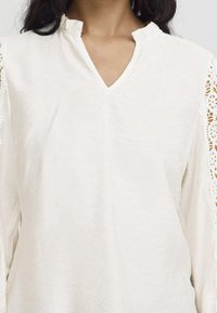 Blouse blanche à manches longues avec un col en V, ornée de délicats accents en dentelle sur les manches et un tissu léger texturé.