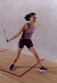 Conjunto atlético en gris y morado oscuro con acentos en rosa; top de tirantes y pantalones cortos ajustados; jugador preparado con una raqueta de squash en una cancha de madera.