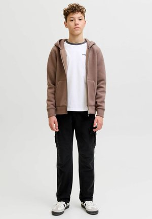 Teismeline poiss seisab, seljas pruun lukuga kapuutsiga dressipluus, valge T-särk, mustad püksid taskutega ning valged tossud mustade triipudega, taust on ühevärviline.