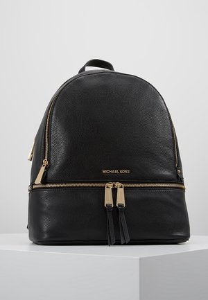 Sort tekstureret læderrygsæk fra Michael Kors med gyldne lynlåse og logo, vist på en hvid piedestal mod en grå baggrund.