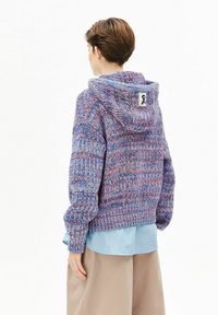Kapuzensweater in Lila und Blau mit strukturierter Oberfläche und geripptem Saum; auf der Rückseite mit einem Etikett versehen und mit einem hellblauen Hemd darunter getragen.