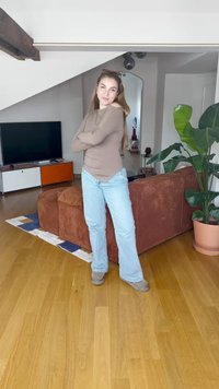 Béžový svetr s dlouhým rukávem, světle modré zvonové džíny a šedé boty, které jsou nošeny v místnosti s dřevěnou podlahou, textilní pohovkou a květináčem.