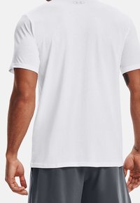 Vit t-shirt med korta ärmar i slät tyg, med rundad urringning och en liten logotyp på övre delen av ryggen. Modellen bär grå shorts.