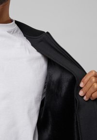 Zwarte zip-up jack met een gladde binnenstof, gedeeltelijk open over een wit t-shirt. Heeft een ribbelkraag en een rits.