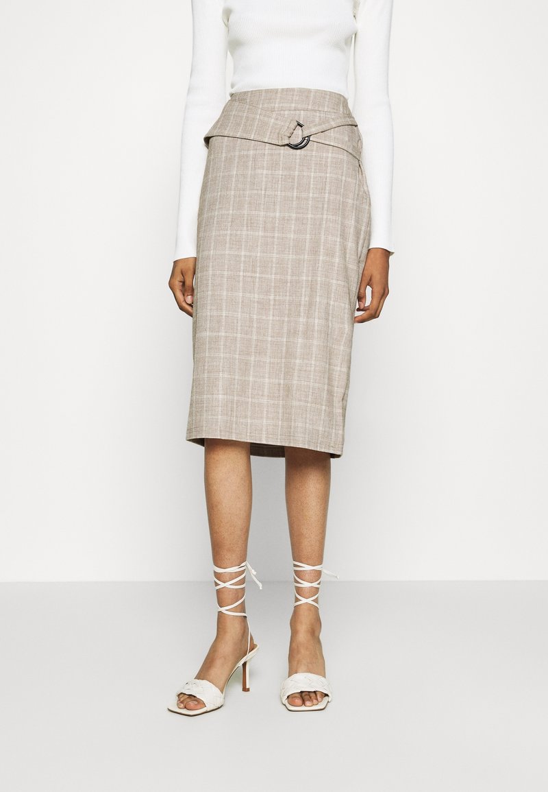 Jupe beige à carreaux longueur genou avec détail asymétrique sur la ceinture, associée à un haut blanc à manches longues et des sandales blanches à talons avec lanières.
