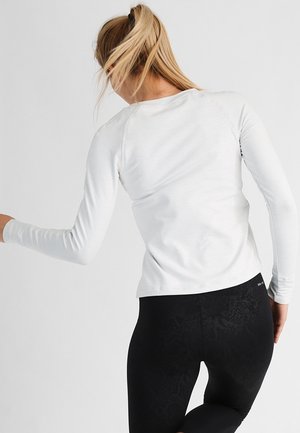 Femme aux cheveux blonds en queue de cheval portant un haut blanc à manches longues et des leggings noirs, se tournant légèrement sur un fond uni.