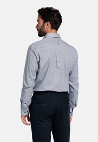 Button-down blouse met een klein blauw-wit geometrisch patroon, lange mouwen en een subtiele rugplooien, gecombineerd met donkere broek.
