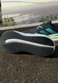 Chaussure de sport noire avec une semelle en caoutchouc texturée, présentant un design épuré, une tige bleu foncé et des accents verts sur le côté.