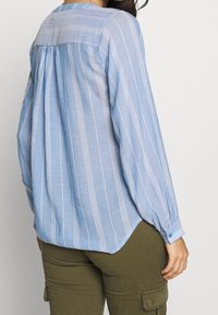 Camicia azzurro chiaro a maniche lunghe con strisce verticali bianche, caratterizzata da un carrè posteriore, orlo arrotondato e polsini con bottoni. Realizzata in un tessuto texturizzato.