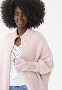 Jasnoróżowy sweter z dzianiny z wysokim kołnierzem, prążkowanymi mankietami i otwartym przodem. Noszony na białej koronkowej bluzce i jasnoniebieskich dżinsach.