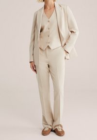 Beige tredelad kostym bestående av en skräddarsydd blazer, väst med fem knappar och åtsittande byxor, kompletterad med bruna sneakers.