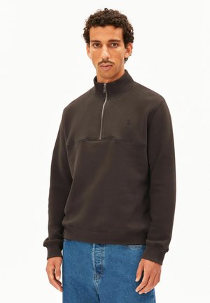 Homme aux cheveux courts et bouclés avec une moustache, portant un sweat-shirt noir à demi-fermeture éclair et un jean bleu, debout devant un fond clair uni.