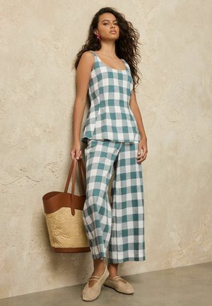 Pikkade juustega naine, kes kannab türkiissinist ja valget ruudulist hõlmiktopi ning pükse, hoides suurt beežikat kootud kotti beeži seina taustal.