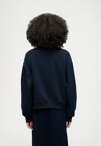 Mörkblå sweatshirt med avslappnad passform, sänkta axlar och ribbade muddar. Tyget verkar mjukt med en slät yta, kort längd.