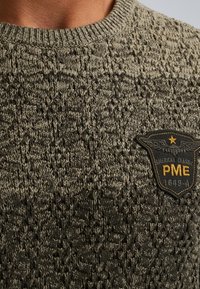 Gebreide trui in donkergroen met een gestructureerd patroon. Heeft een geel en zwart embleem met "PME 1649" op de linkerkant van de borst.