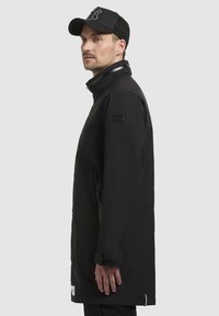 Chaqueta impermeable negra con un cuello alto, bolsillos con cremallera, aberturas laterales y un parche con el logo en la manga. Llevada con una gorra de béisbol negra.