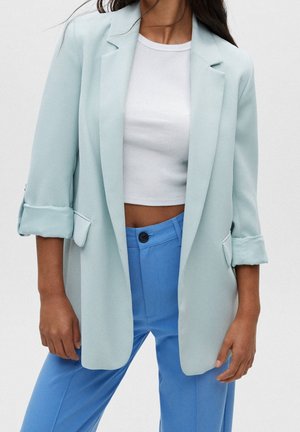 Frau trägt einen hellblauen Blazer mit hochgekrempelten Ärmeln, ein weißes Crop-Top und leuchtend blaue High-Waist-Hosen vor einem schlichten Hintergrund.