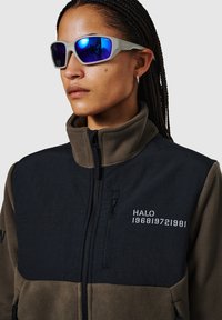 Kurtka polarowa w ciemnej zieleni i czerni, z wysokim kołnierzem, zapinana na zamek kieszonka z przodu oraz haftowanym napisem "HALO 196819721981." Okulary przeciwsłoneczne z niebieskimi soczewkami.