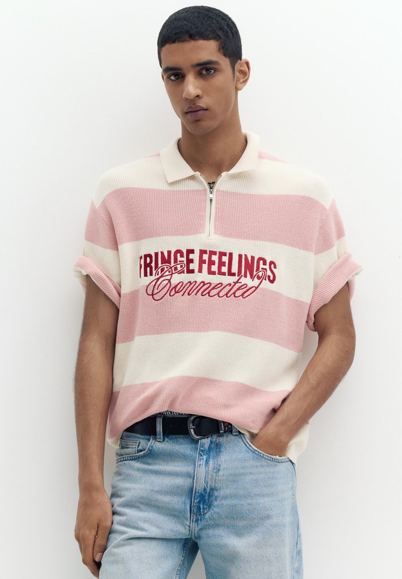 Jeune homme aux cheveux courts porte un pull à manches courtes rayé rose et blanc avec le texte « Fringe Feelings Connected » et un jean bleu clair.