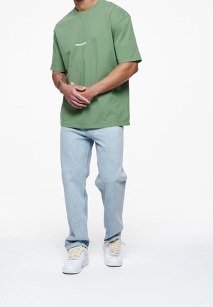 Hombre con camiseta verde, jeans azul claros y zapatillas blancas con cordones gruesos color beige, de pie con las manos entrelazadas frente a un fondo blanco.