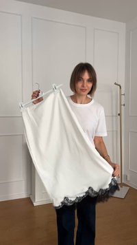 Weißer Rock mit schwarzem Spitzenbesatz, glatte Textur, gerade Form, an einem Kleiderbügel aufgehängt. Begleitet von einem schlichten weißen Hemd und dunklen Hosen.