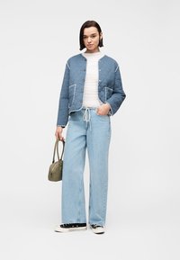 Νεαρή γυναίκα που φοράει μπλε μπουφάν με καπιτονέ ύφασμα, λευκό τοπ, ανοιχτό μπλε τζιν με φαρδιά πόδια, μαύρα αθλητικά παπούτσια, κρατώντας μικρή ελιά τσάντα, όρθια σε λευκό φόντο.